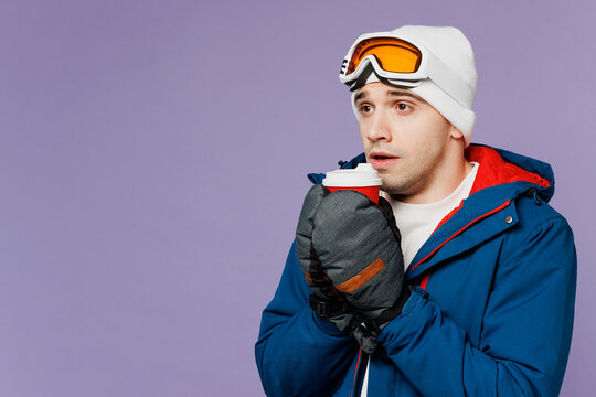 Skier sad man wear warm blue windbreaker jacket ski goggles mask mittens hold takeaway delivery cup coffee to go spend extreme weekend winter season in mountains isolated on plain purple background.