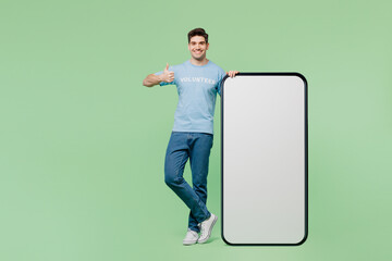 Full body young fun man wear blue t-shirt title volunteer big huge blank screen area mobile cell phone show thumb up isolated on plain green background. Voluntary free work help charity grace concept.