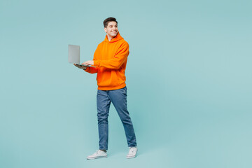 Full body side view young IT man he wears orange hoody casual clothes hold use work on laptop pc...