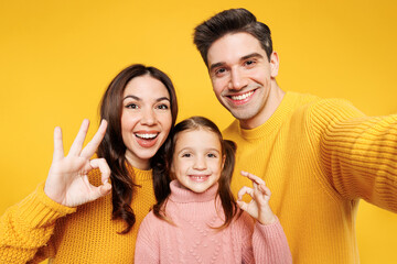 Fototapeta premium Close up young parents mom dad with child kid girl 7-8 years old wear pink sweater casual clothes do selfie shot mobile cell phone show ok okay isolated on plain yellow background. Family day concept.