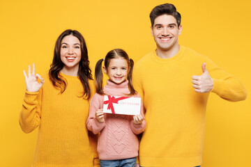 Obraz na płótnie Canvas Young parents mom dad with child kid girl 7-8 years old wear pink sweater casual clothes hold gift coupon voucher card for store show thumb up isolated on plain yellow background. Family day concept.