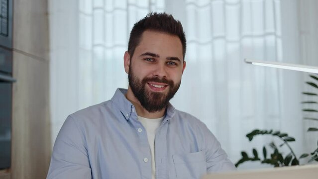Portrait of caucasian man smiling and looking at camera while sitting at home desk with modern laptop. Bearded male freelancer in casual wear expressing satisfaction from favorite remote work.
