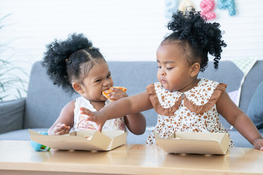 Cute African children eating pizza at home. Kids enjoy and having fun with tasty lunch meal together. Happy little girls sharing yummy pizza