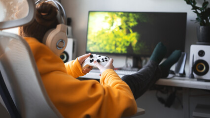 Teenage girl with headset is sitting in a comfortable computer chair, holding a white gamepad in her hands and playing a game on a PC or console.  © Aboltin