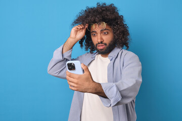Young surprised curly Arabian man lifts glasses from eyes and looks at mobile phone screen feeling...