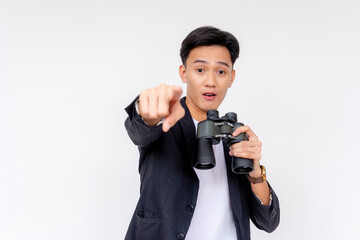 Amazed young asian man in a suit points directly at camera while holding binoculars on a white background