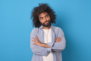 Young cunning bearded Arabian man with crossed arms looks to side pondering plan of revenge for offender or developing strategy for seducing new girlfriend stands posing on blue background. - obrazy, fototapety, plakaty