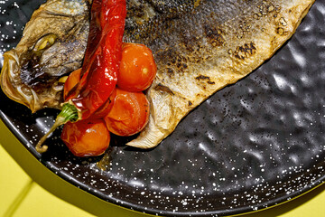 Fried sea bass with baked tomatoes in a black plate on a yellow background