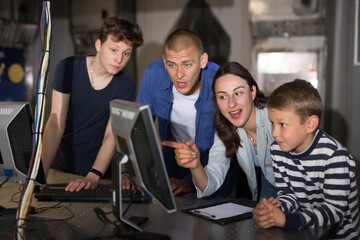 Cheerful young parents with two sons spending funny time in quest room decorated as lost underground shelter using computer to find solution to puzzle