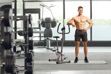Fitness coach posing shirtless and leaning on an exercise bike at a gym