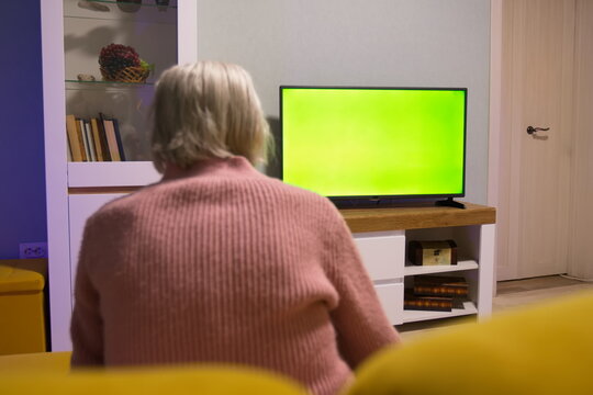 Grandma is watching TV. Green screen. Grandma is sitting on the sofa at home. In front of her is a green screen TV. 