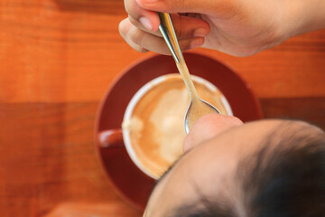 A woman holds a spoon and scoops hot coffee cream foam into her mouth on the table.