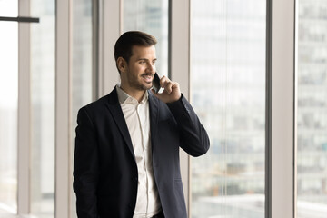 Positive confident handsome businessman talking on mobile phone at large office window, looking away at city view, thinking on future project success, discussing work plan on cellphone call