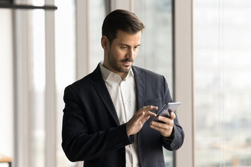 Serious handsome businessman in suit typing message on smartphone, using work online application on mobile phone, browsing professional social network, website on Internet