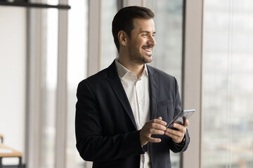 Fototapeta premium Happy confident entrepreneur man using mobile phone for business online communication, holding smartphone, looking at window away, thinking on wireless technology, smiling
