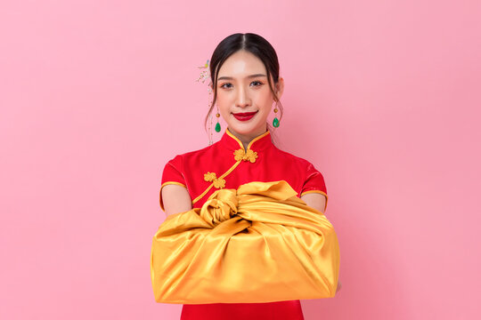 Chinese new year portrait of elegant Asian woman in traditional cheongsam dress giving golden gift box in pink color background
