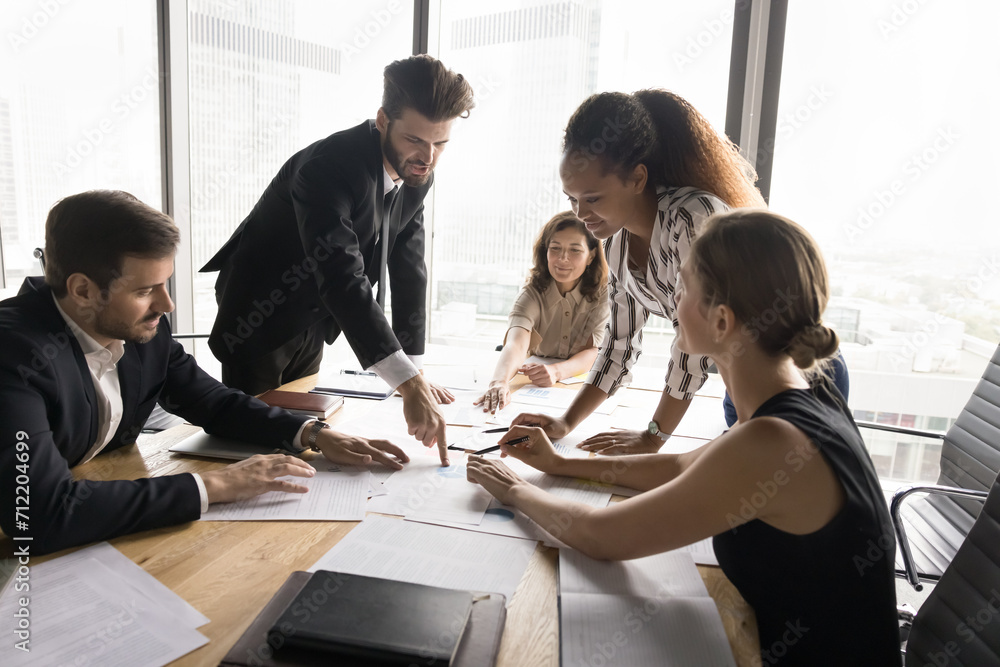 Poster Busy marketing team analyzing work results together, discussing paper sales reports, bending over meeting table, pointing at documents, speaking, brainstorming on creative strategy