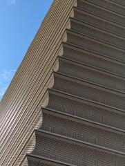 A part of a building with an intricate and repetitive pattern on its exterior. The building has multiple layers, each protruding outward, creating a step-like structure on the facade.