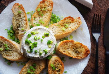 Brie cheese with green onions with bread croutons on a plate next to cutlery on a dark background