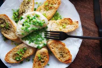 Brie cheese with green onions with bread croutons on a plate next to cutlery on a dark background