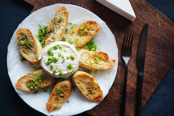 Brie cheese with green onions with bread croutons on a plate next to cutlery on a dark background