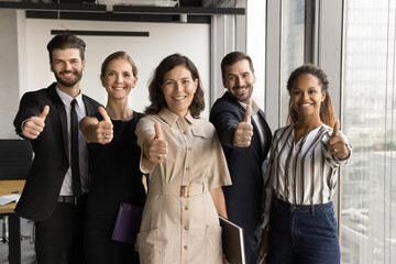 Happy diverse team and mature female leader posing for portrait in office space, showing thumb up...