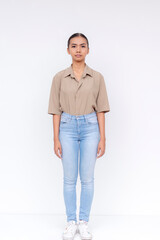 Fototapeta premium Portrait of a confident young Filipino woman in a khaki blouse and light blue jeans standing with a neutral expression against a white backdrop.