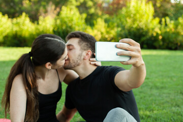 Pareja heterosexual caucasica enamorados haciendose un selfie en el cesped del parque posando besándose - obrazy, fototapety, plakaty
