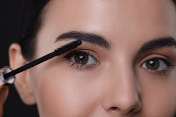 Beautiful young woman applying mascara on black background, closeup