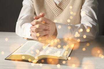 Woman praying over Bible at white wooden table, closeup. Bokeh effect