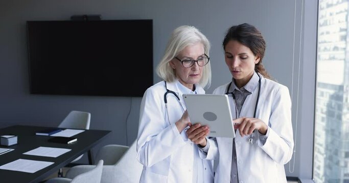 Two serious younger and elder doctor women in uniform coats using tablet together, discussing online content, job medical application, meeting, talking. Practitioner consulting colleague for expertise