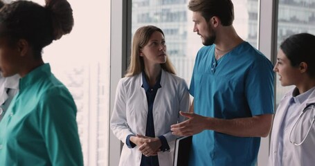 Team of serious diverse medical colleagues in white and blue uniforms walking together past camera in hospital office hall, talking, discussing difficult job cases, career in medicine, teamwork