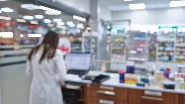 Defocused view of drugstore with a pharmacist working at the seller counter cash register in front of computer. Pharmacy stocked shelves with medicines, healthcare industry blurred background
