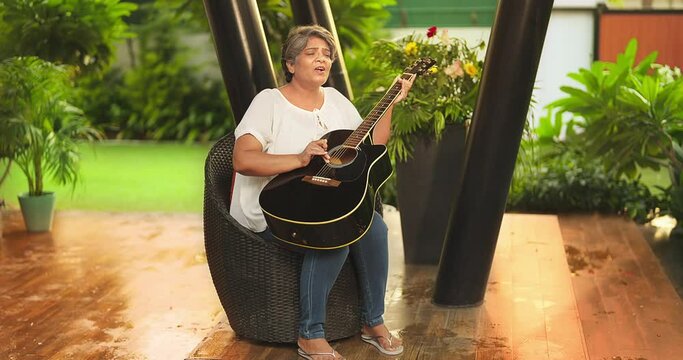 Asian Indian Hindu happy older middle aged woman lady sitting chair singing song play guitar enjoy fun joy pastime outdoor home park smiling old elder adult female tuning music lyric house garden