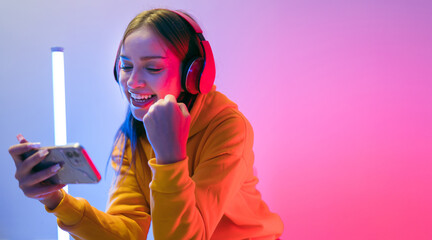 A young Caucasian beautiful woman in a yellow hoodie playing a video game using a smartphone and headphones with fun, focus, surprise, and a winning victory face with blue and red neon light.
