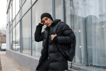 fashion man with a hat in a black stylish down jacket with a backpack stands near a glass building...