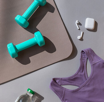 Sport or workout concept flat lay with blue dumbbells, sport wear, yoga mat and white wireless headphones on the grey background.