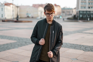 Stylish urban handsome guy with glasses with a hairstyle in a fashion black jacket walks in the city