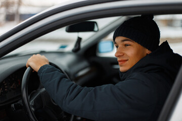 Happy man with a winter knitted hat and jacket sits in a car on a winter snow day. Frost and transport. Engine starting