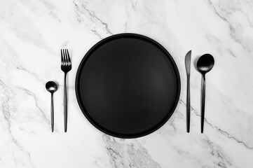 Top view of black cutlery and black plate on white marble background. Creative table setting flat...