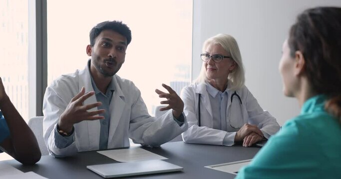 Happy young Indian medical professional man in white coat discussing occupation with doctor colleagues at meeting table, talking to diverse coworkers in uniform, smiling, laughing