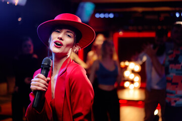 Joy and music. Happy young woman in red hat is singing in karaoke nightclub - obrazy, fototapety, plakaty