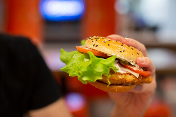Hands holding a hamburger sprinkled with sesame seeds on a blurred background.