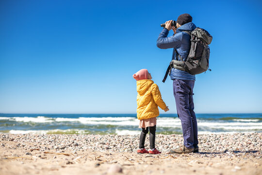 A tourist man standing on the seashore with his little daughter, using binoculars to observe the coastal landscape