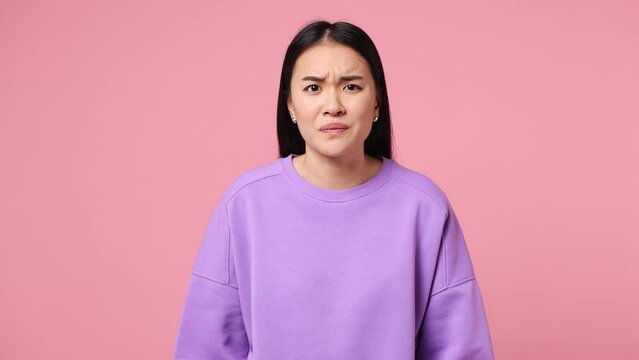 Sad upset worried young woman of Asian ethnicity wear purple sweatshirt ask what hears fake news propaganda has some problems gesticulating with hands isolated on plain pastel light pink background
