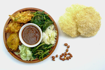 Pecel or pecal is traditional Javanese salad with peanut sauce. Served on a wooden plate with bakwan or vegetable fritters and fried chicken isolated on white background