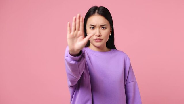 Serious strict severe young woman of Asian ethnicity wearing purple sweatshirt say no hold palm folded crossed hands in stop gesture isolated on plain pastel light pink background. Lifestyle concept