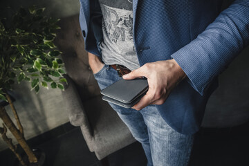 A man in a blue jacket holds a gray leather wallet in his hand against the background of the interior. The atmosphere of comfort and business confidence emphasizes the status and fashionable style