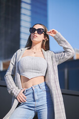 Dark haired woman with fit body wears sunglasses while posing artistically under the sunlight between modern buildings. She wears a top, blazer and jeans.