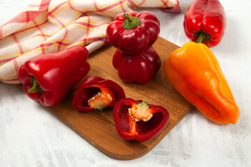 Cutting board with several whole and halves of red and yellow bell pepper and red kitchen towel on white wooden background. .